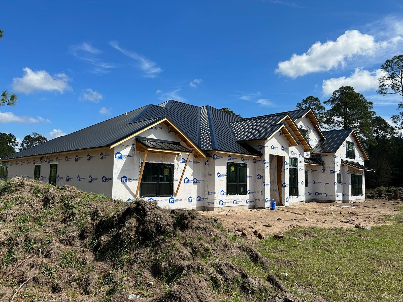 House under construction with blue wrap, unfinished windows, and black roof. Green grass and blue sky.