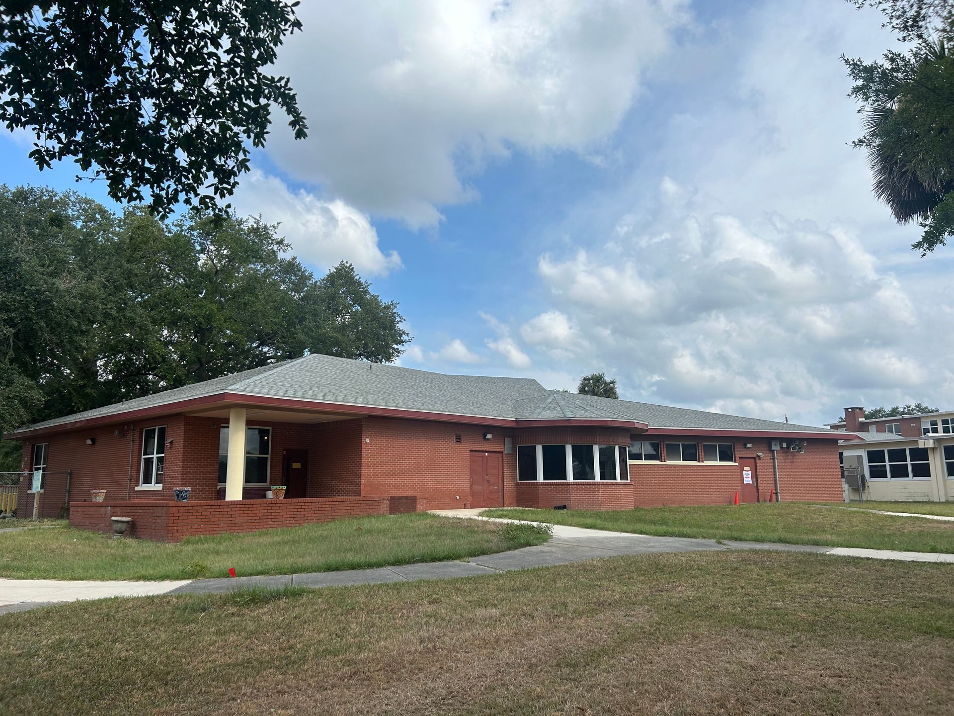 Red brick building with a gray roof under a partly cloudy sky. Green grass in front, trees in the background.