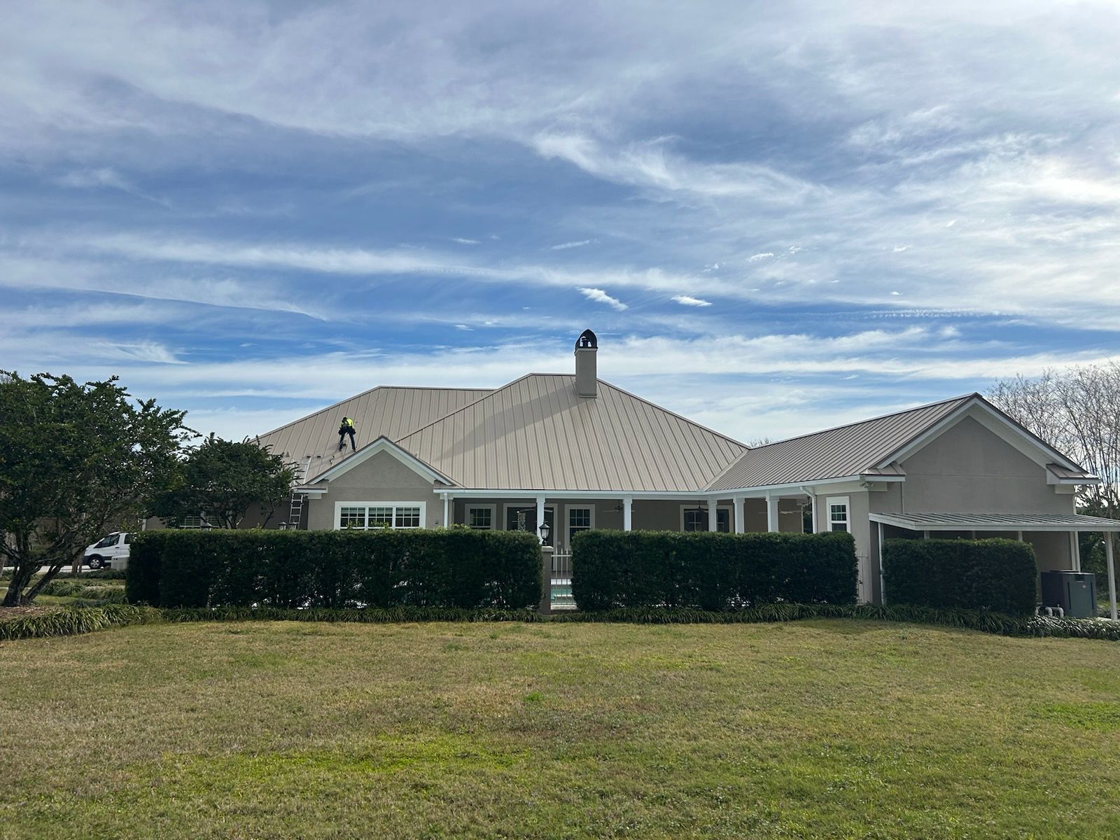 Beige house with a brown roof and a green hedge row in front of a blue sky.