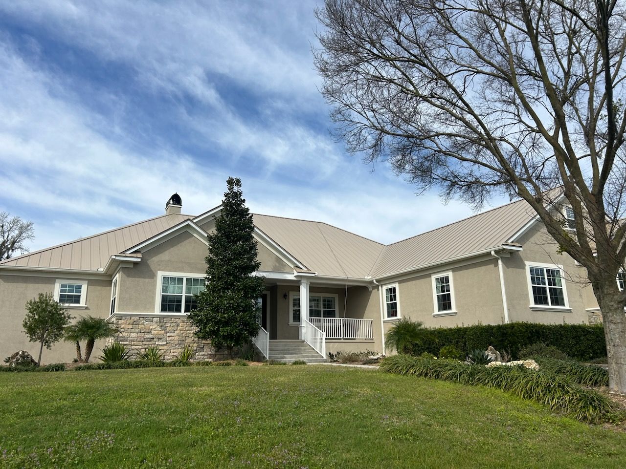 Tan ranch-style house with metal roof, stone accents, and green lawn under a blue sky.