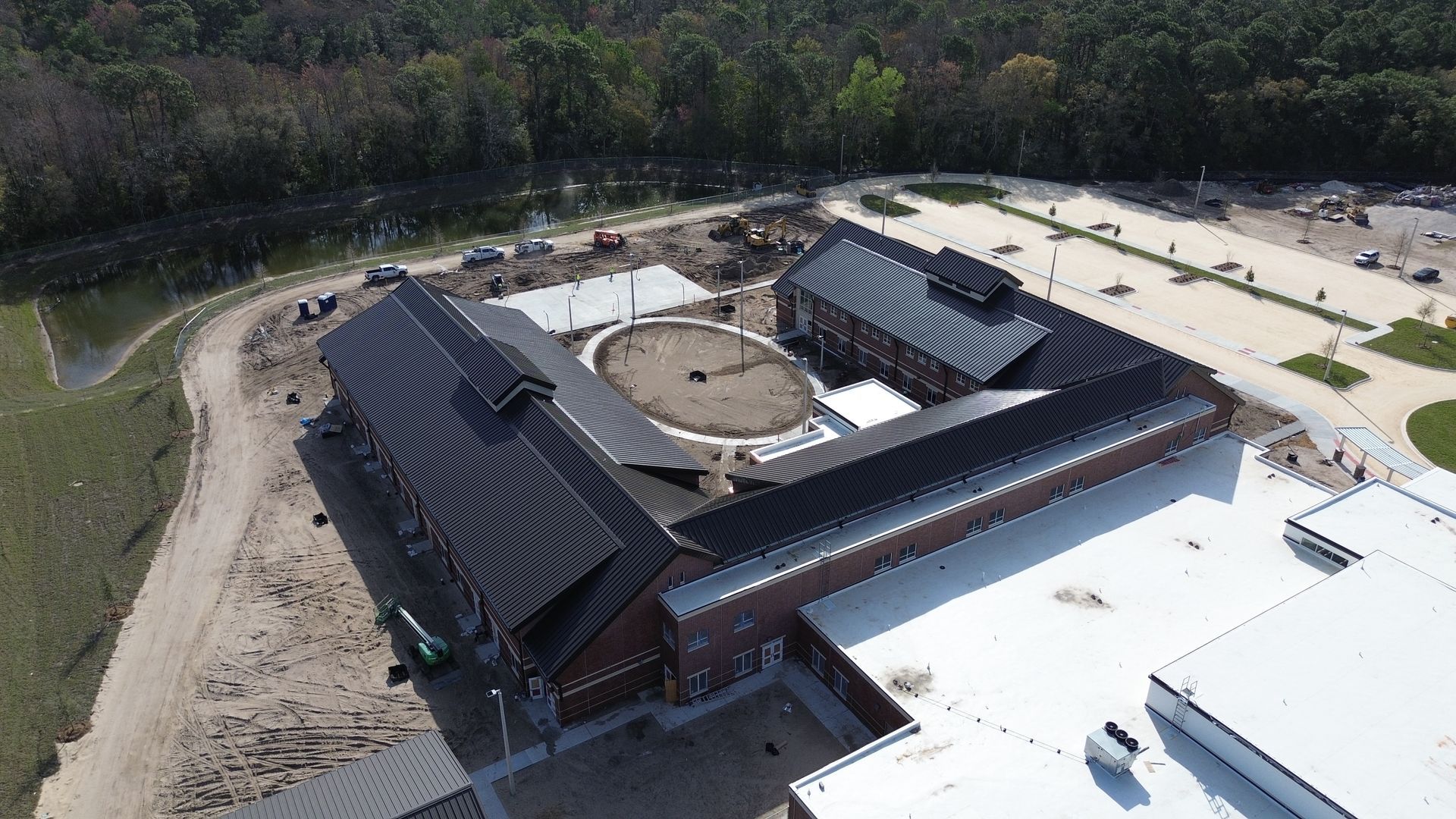 Aerial view of a brick building with a dark roof and a central circular area. Construction site, trees in background.