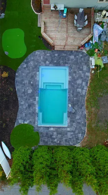 Overhead view of a swimming pool with blue water surrounded by dark gray paving stones. Green grass and putting green nearby.