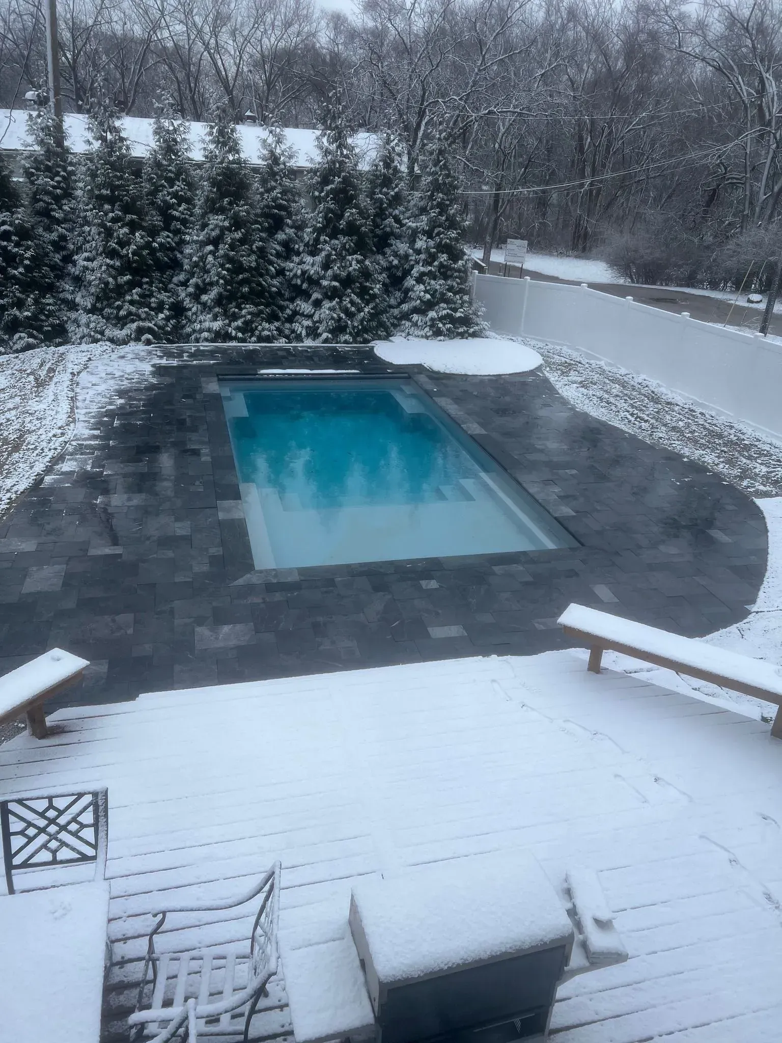 Snowy backyard with a rectangular pool, dark pavers, and evergreen trees. Two benches covered in snow.
