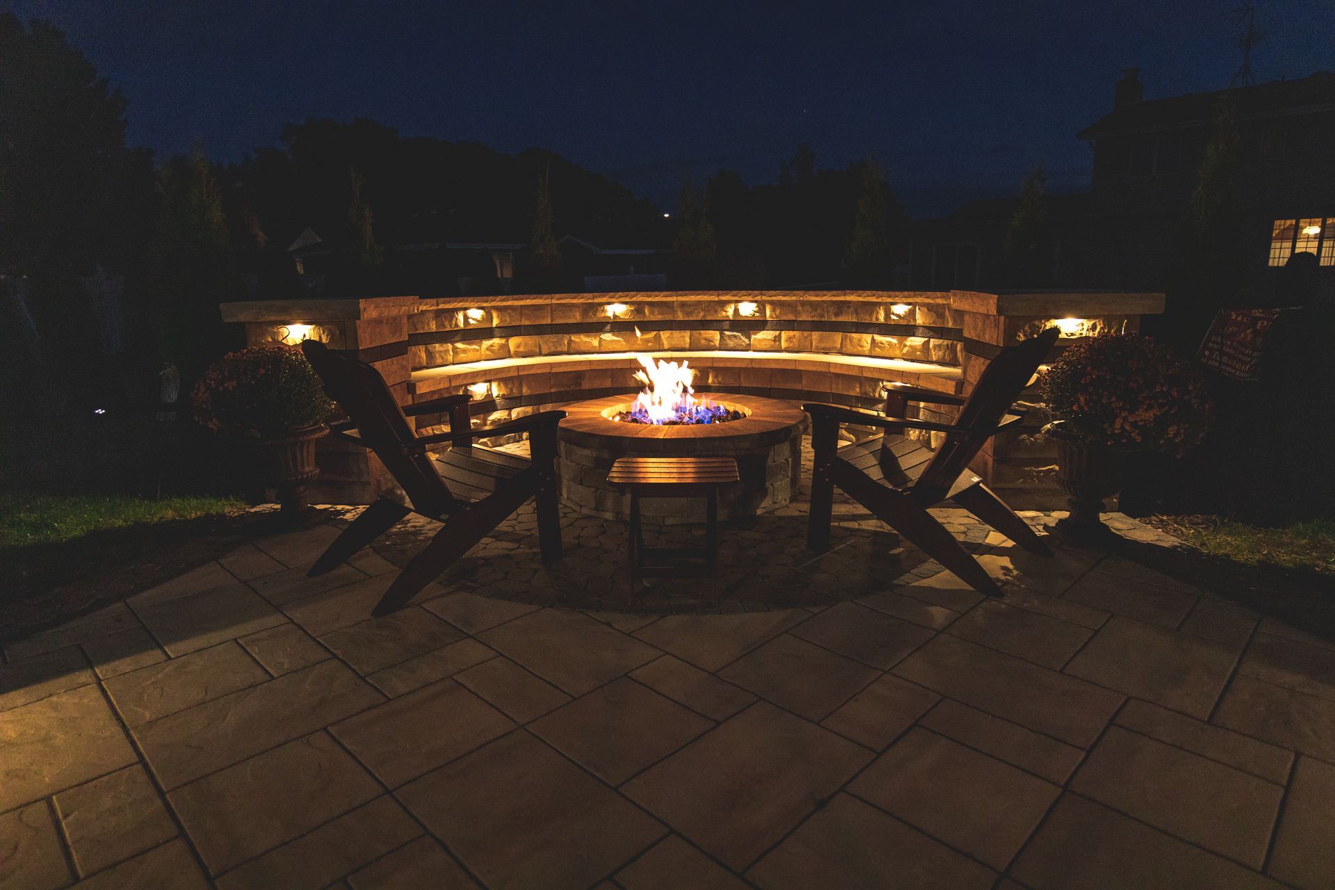 Stone fire pit with built-in seating lit up at night with two chairs in the foreground.