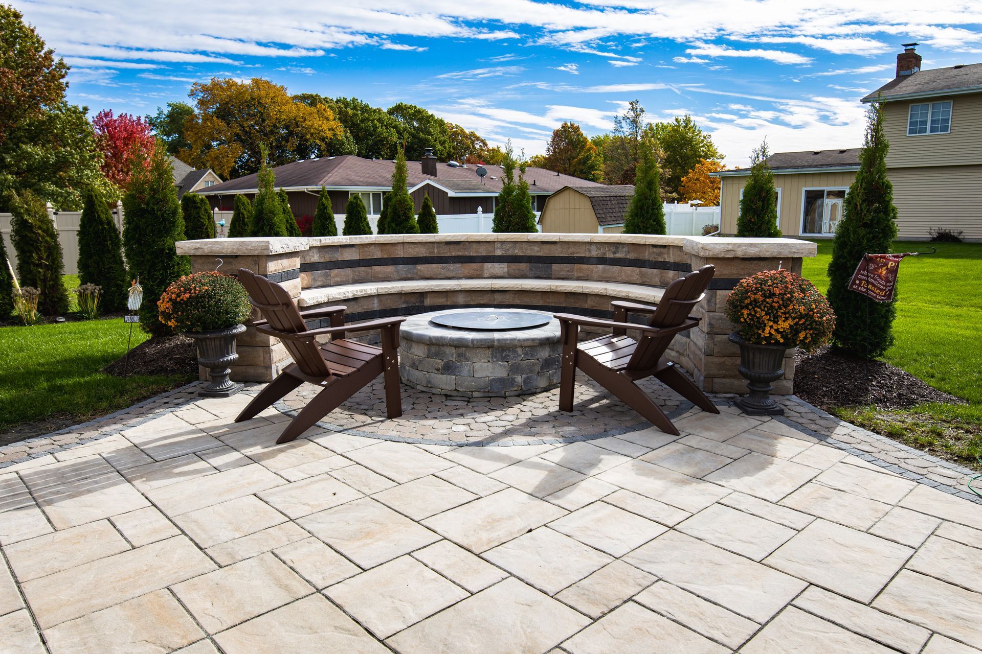 Patio with fire pit, stone seating, and two wooden chairs. Green lawn, trees, and blue sky in background.