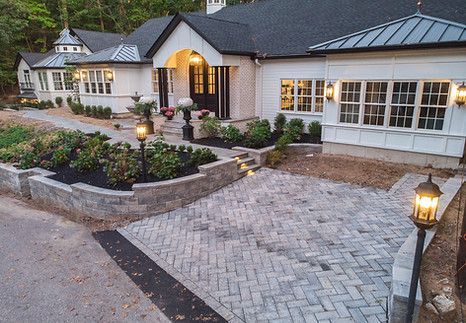 White house with brick and dark roof, gray paver driveway, lampposts, and landscaping.