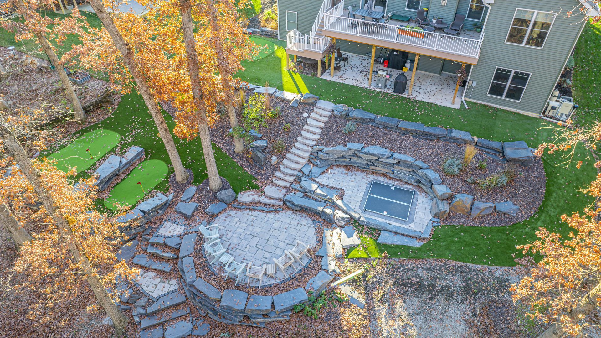 Aerial view of a backyard with a fire pit, hot tub, putting greens, stairs, and a house with a deck.