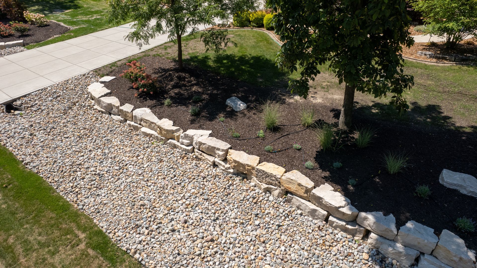 Stone retaining wall bordering a gravel area and a landscaped garden bed with trees.