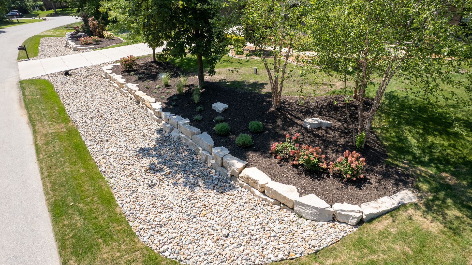 Landscaped yard with white stone bed and retaining wall, dark mulch, trees, and plants.
