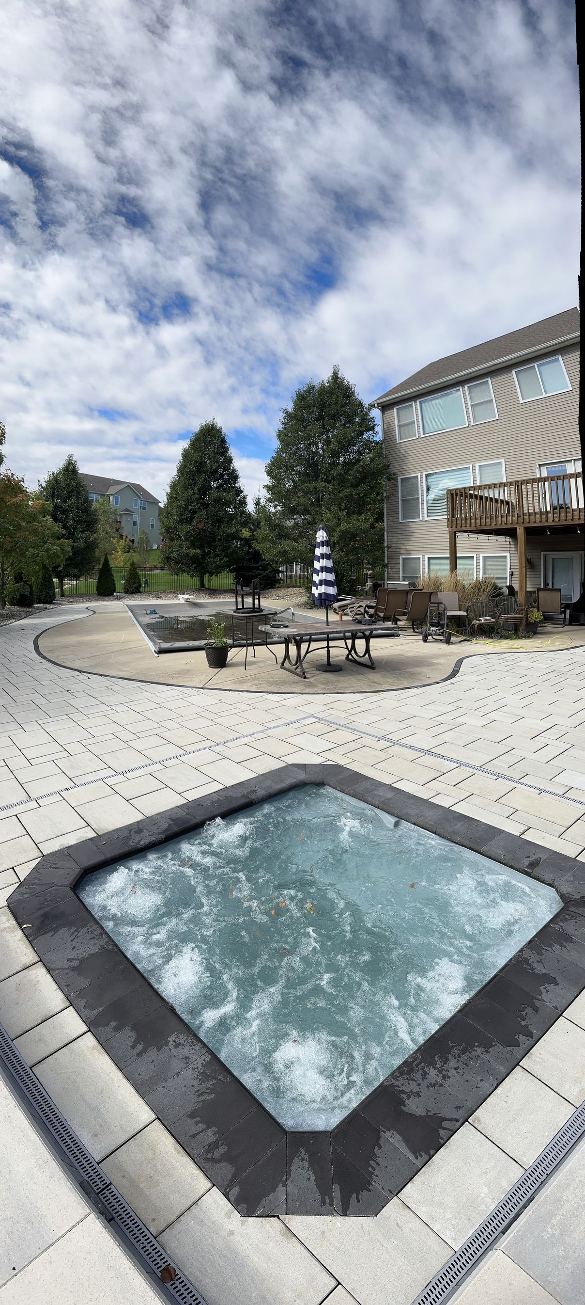 Bubbling hot tub in paved backyard, trees, blue sky.