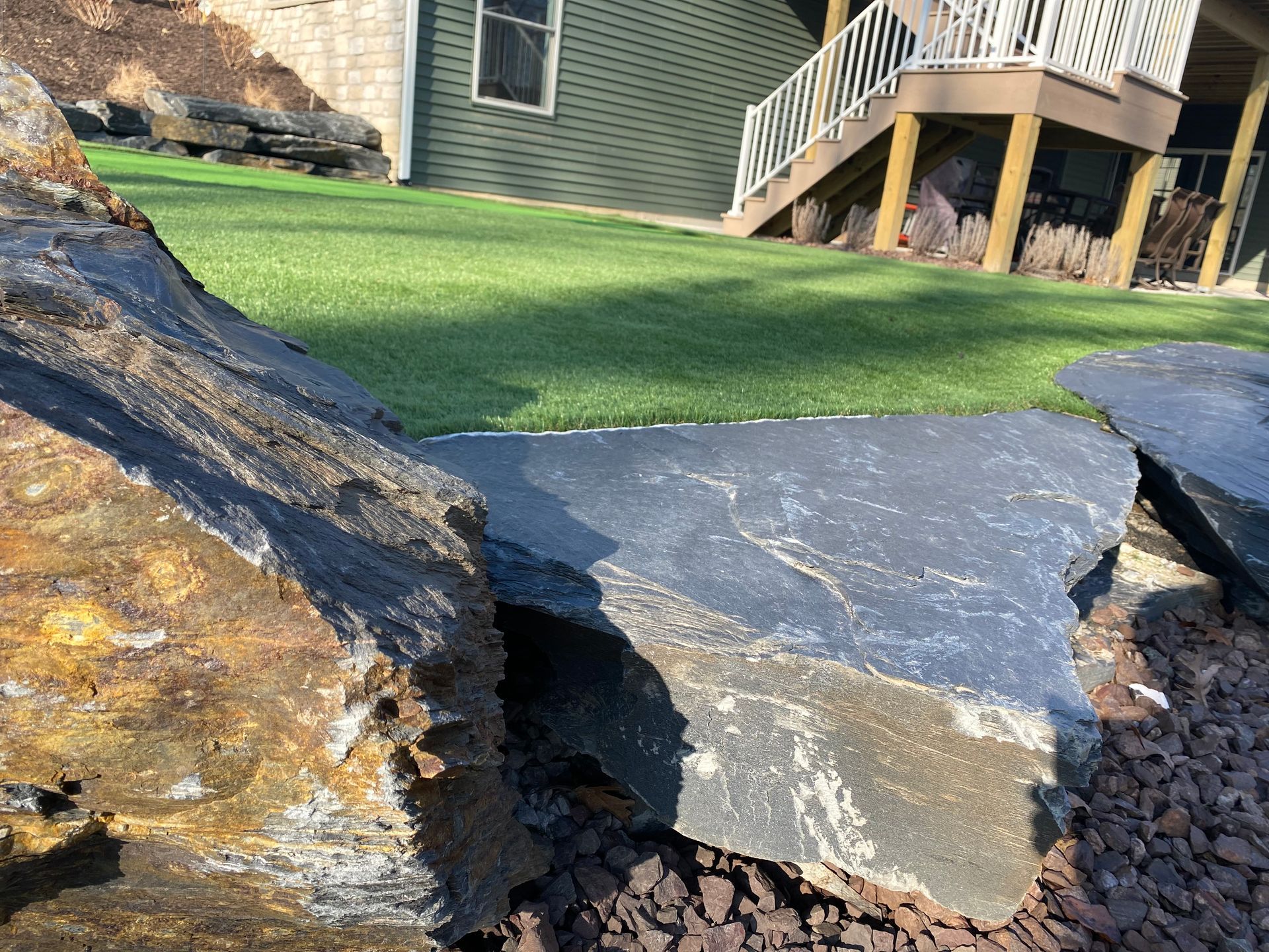 Large dark rocks bordering green artificial turf with a green house and deck in the background.