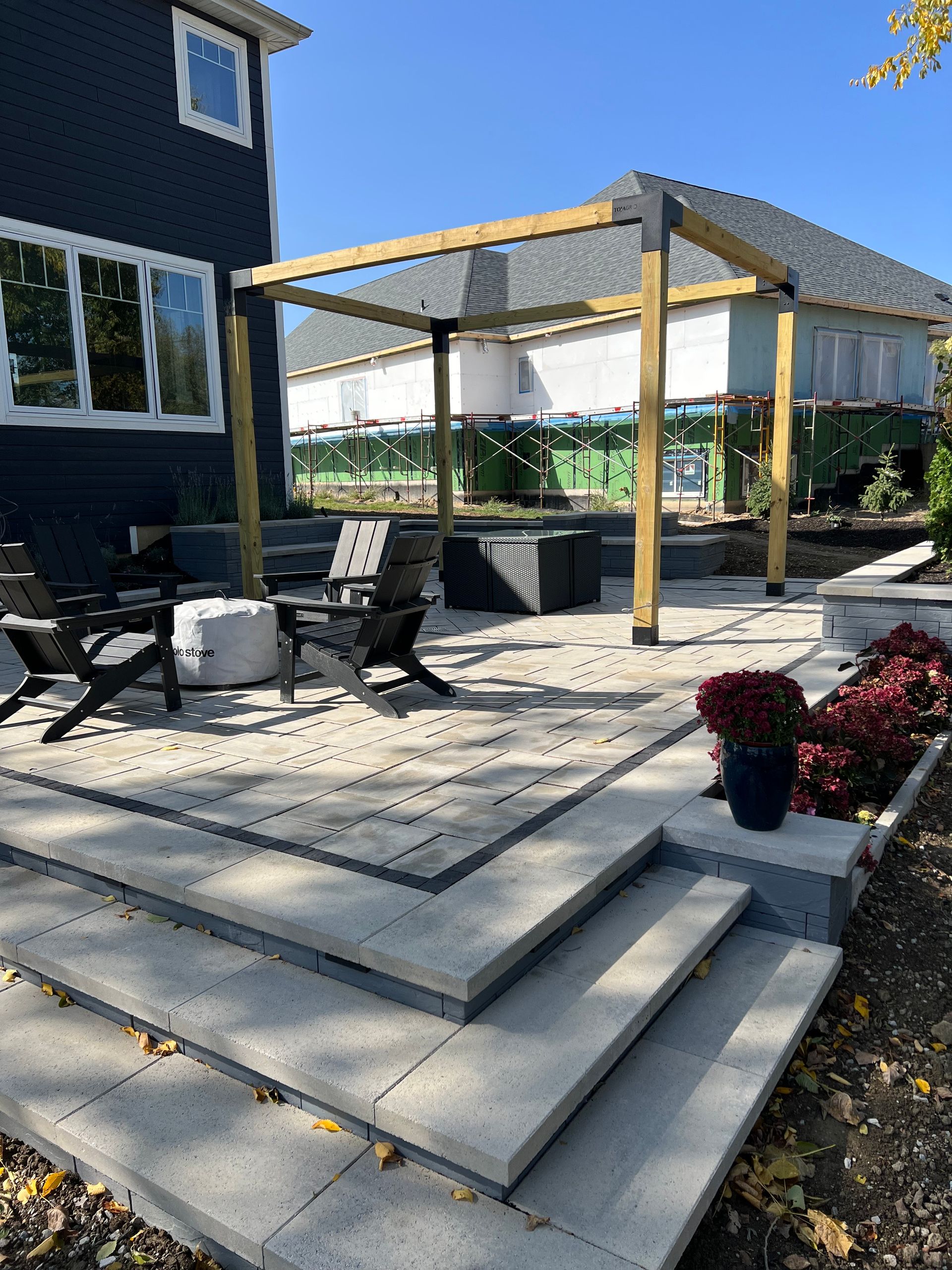 Stone patio with steps and pergola, next to a house. Outdoor furniture and plants present.