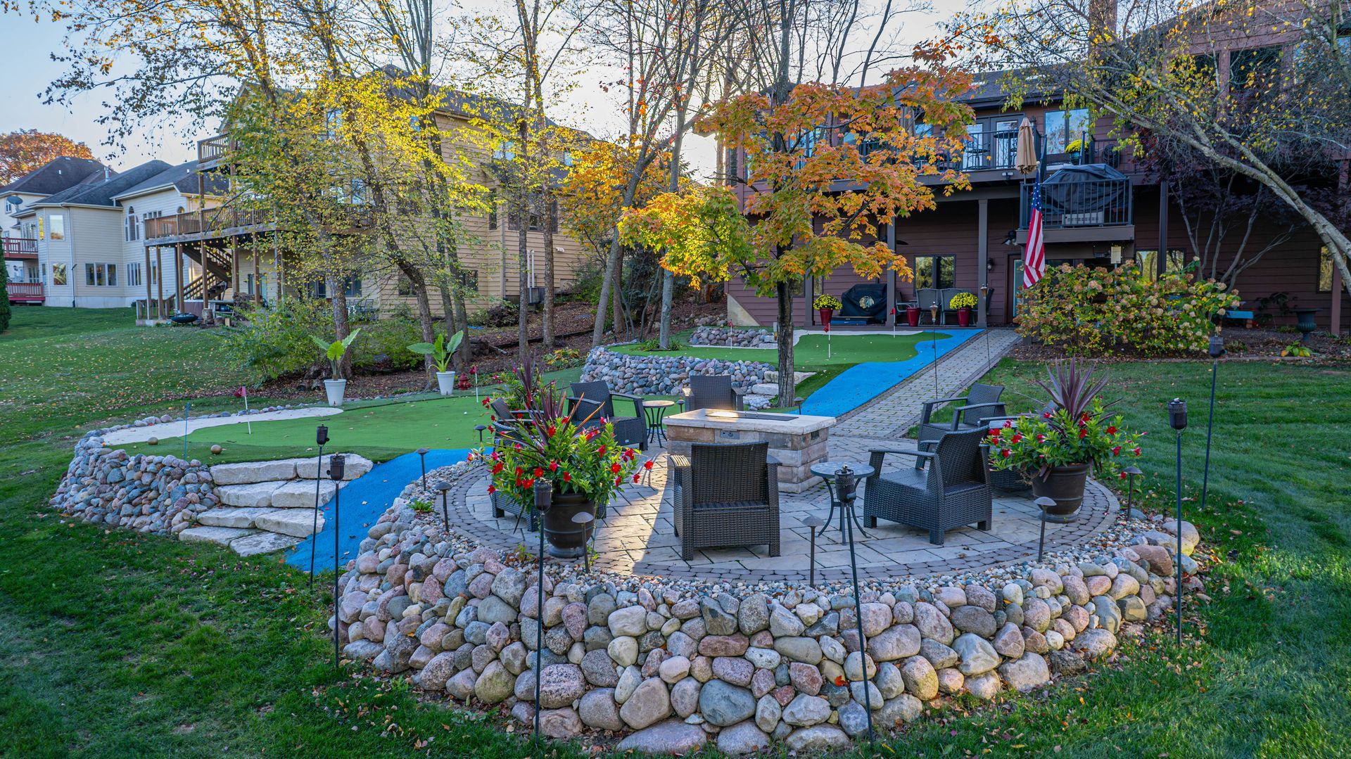A backyard with a stone patio, fire pit, and a blue path leading to a putting green.