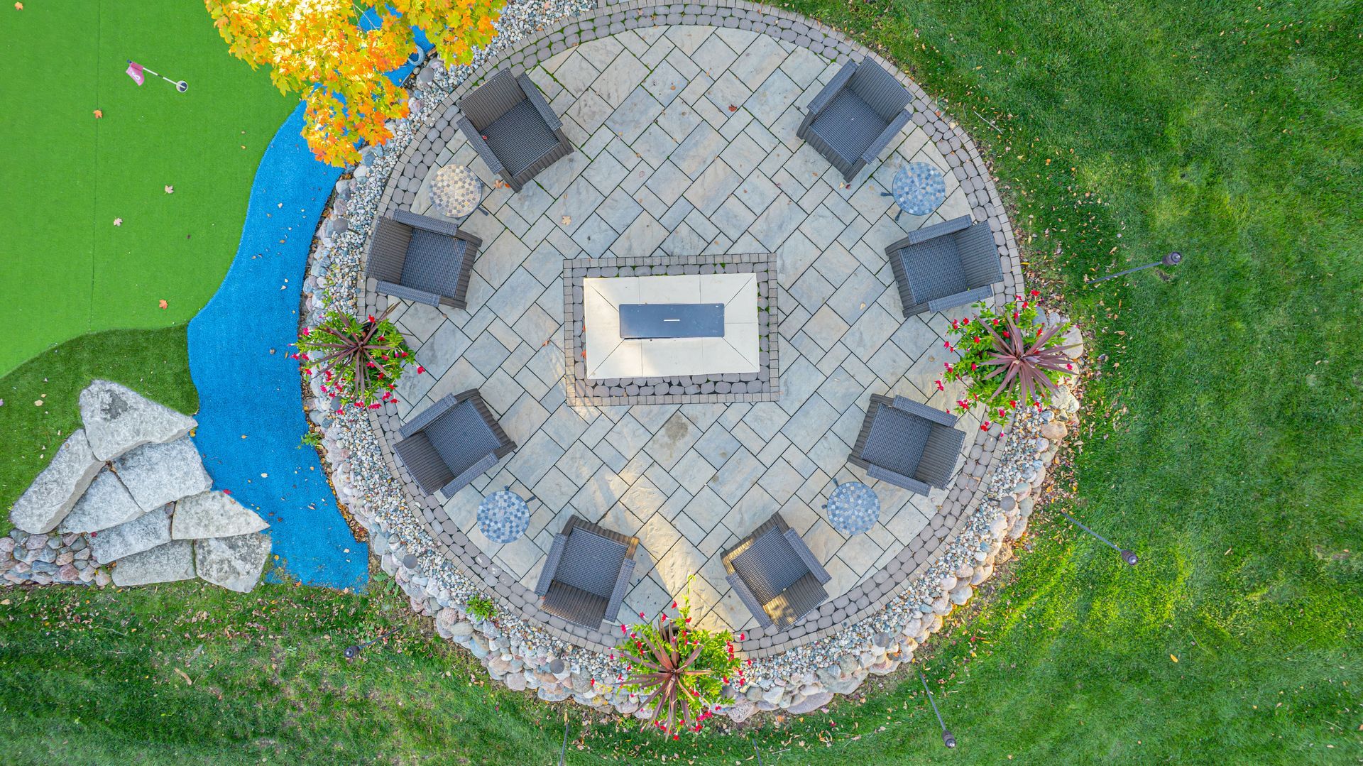 Overhead view of a circular stone patio with eight chairs around a rectangular fire pit. 