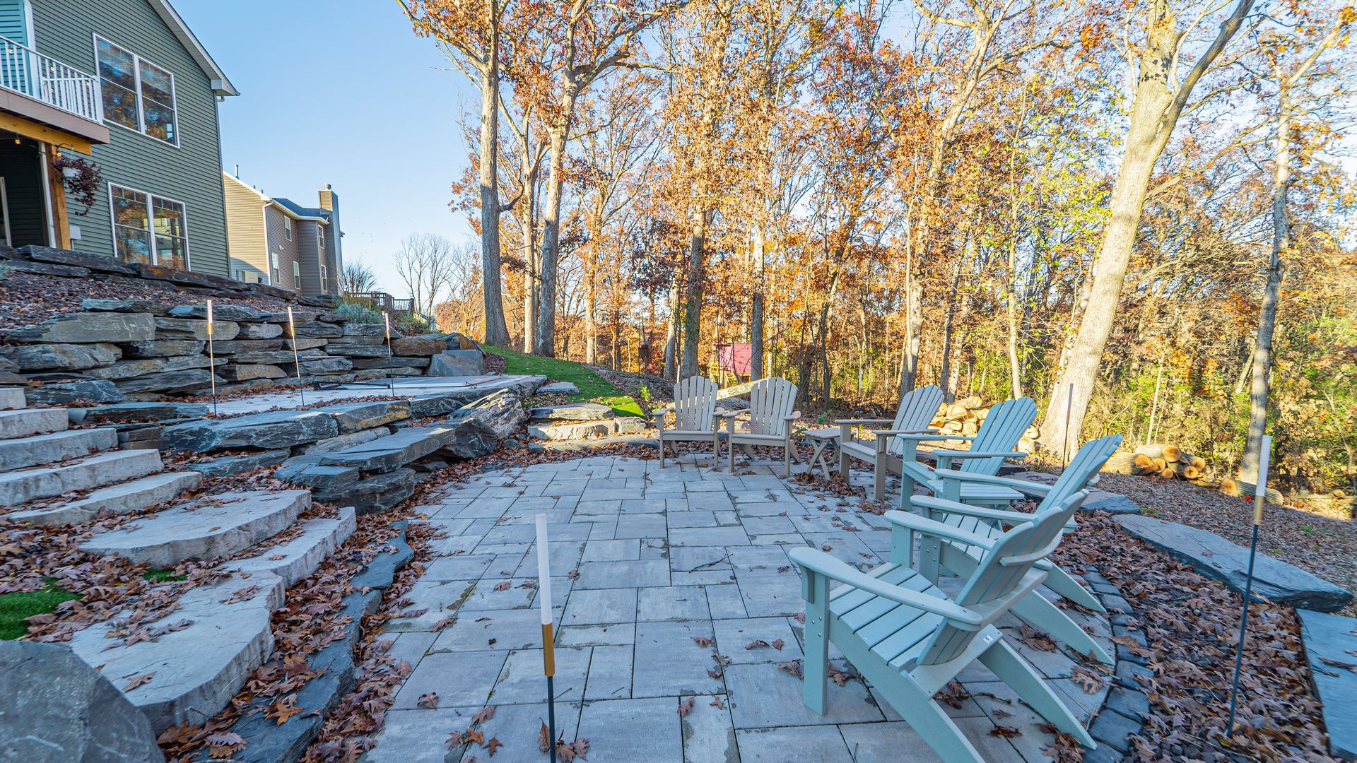 Stone patio with Adirondack chairs, near a terraced yard and a house with trees.