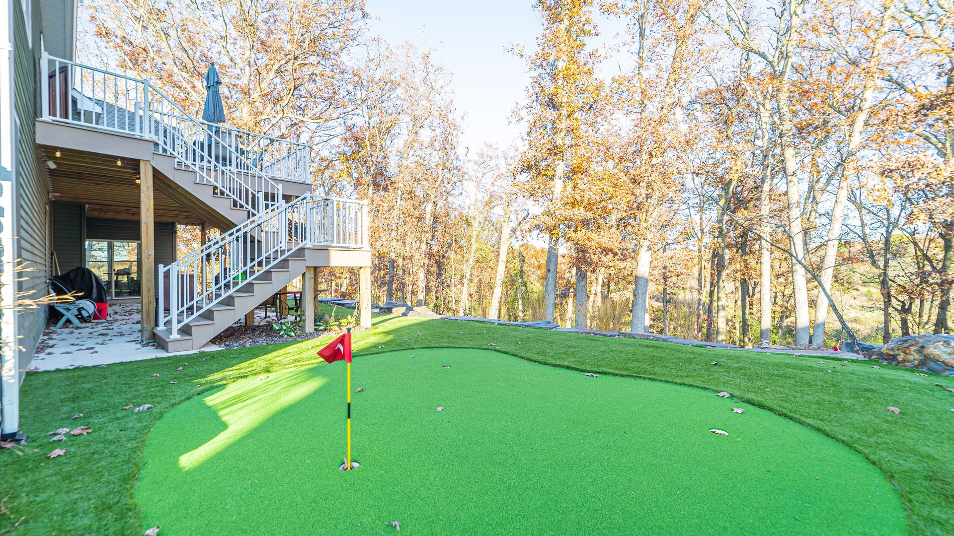 Backyard with a green putting area, a flag, and a deck with stairs, surrounded by trees.