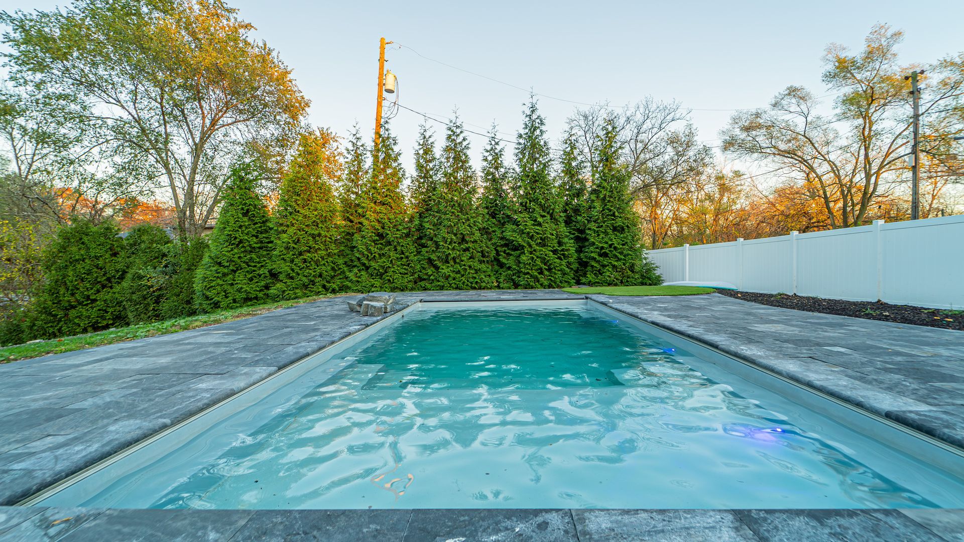Swimming pool with turquoise water, surrounded by stone and green trees, with a white fence in the background.