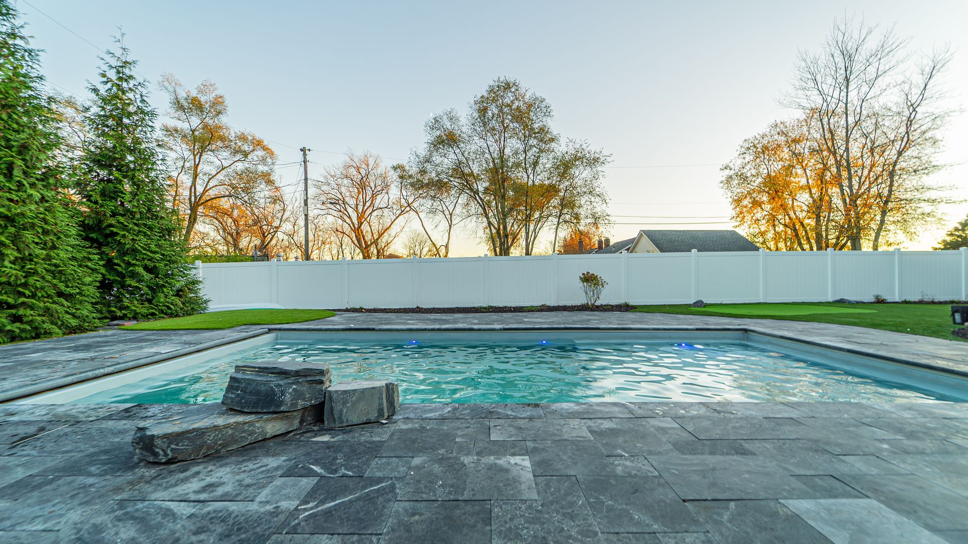Backyard pool with stone patio and white fence; trees in background.