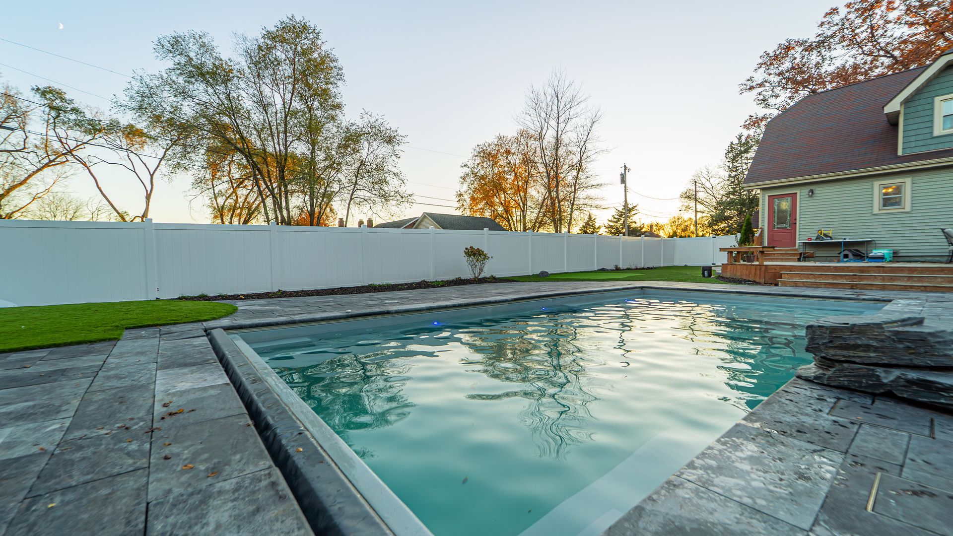Swimming pool with stone patio, green lawn, white fence, and house with a red door.