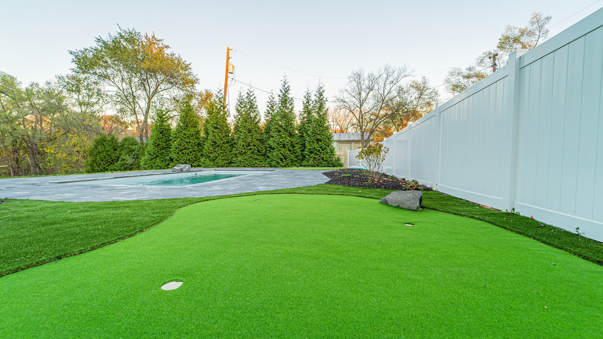 Backyard with green artificial turf, small putting green, pool, and white fence.