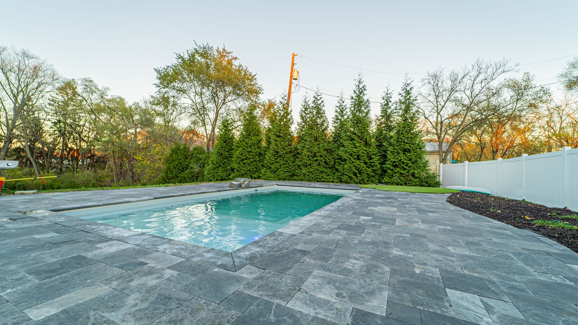 Backyard with rectangular pool, gray paving stones, green trees, white fence.