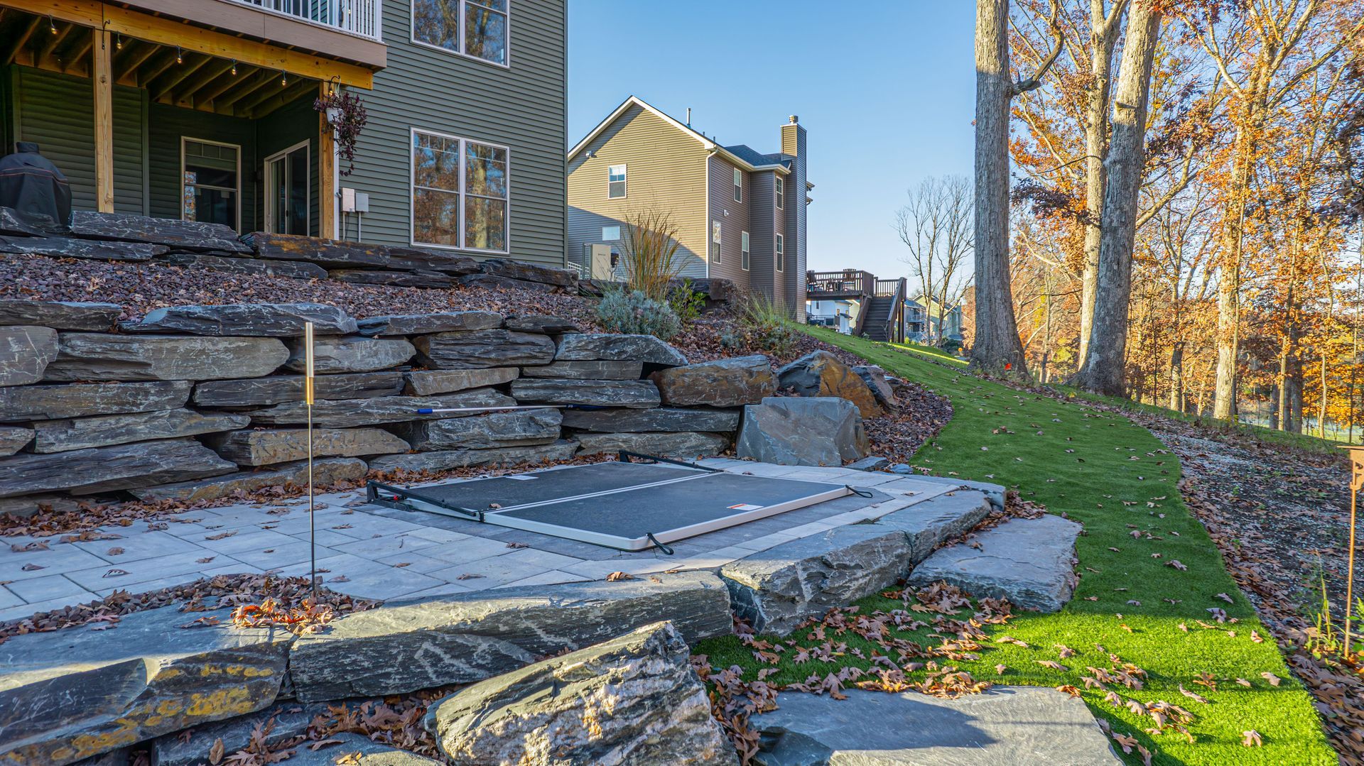 Stone patio with hot tub cover, retaining wall, and green lawn. Houses in background.