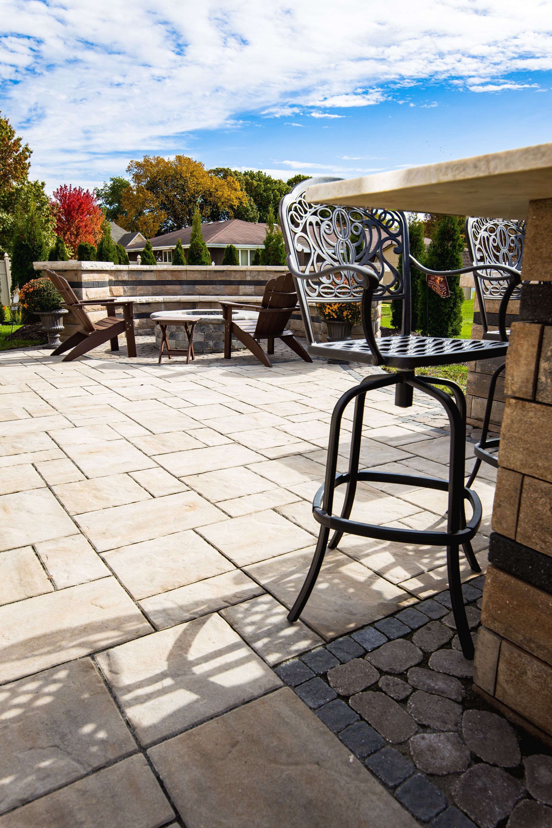 Patio with stone pavers, bar seating, and wooden seating. Bright blue sky with trees in the background.