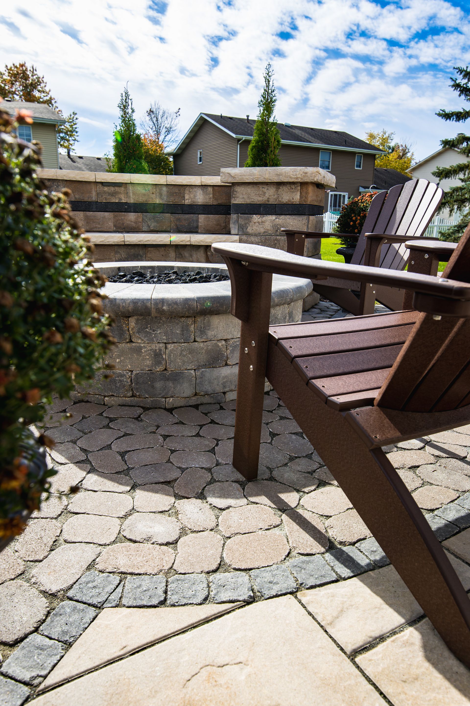 Stone fire pit with brown Adirondack chairs on a paved patio, residential setting.