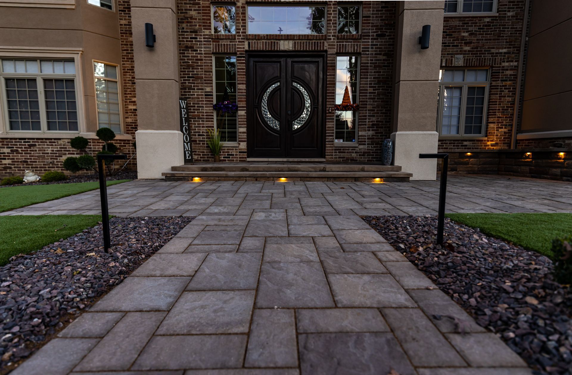 Brick walkway leads to a house with double doors. The walkway is lined with small black posts and decorative stones.