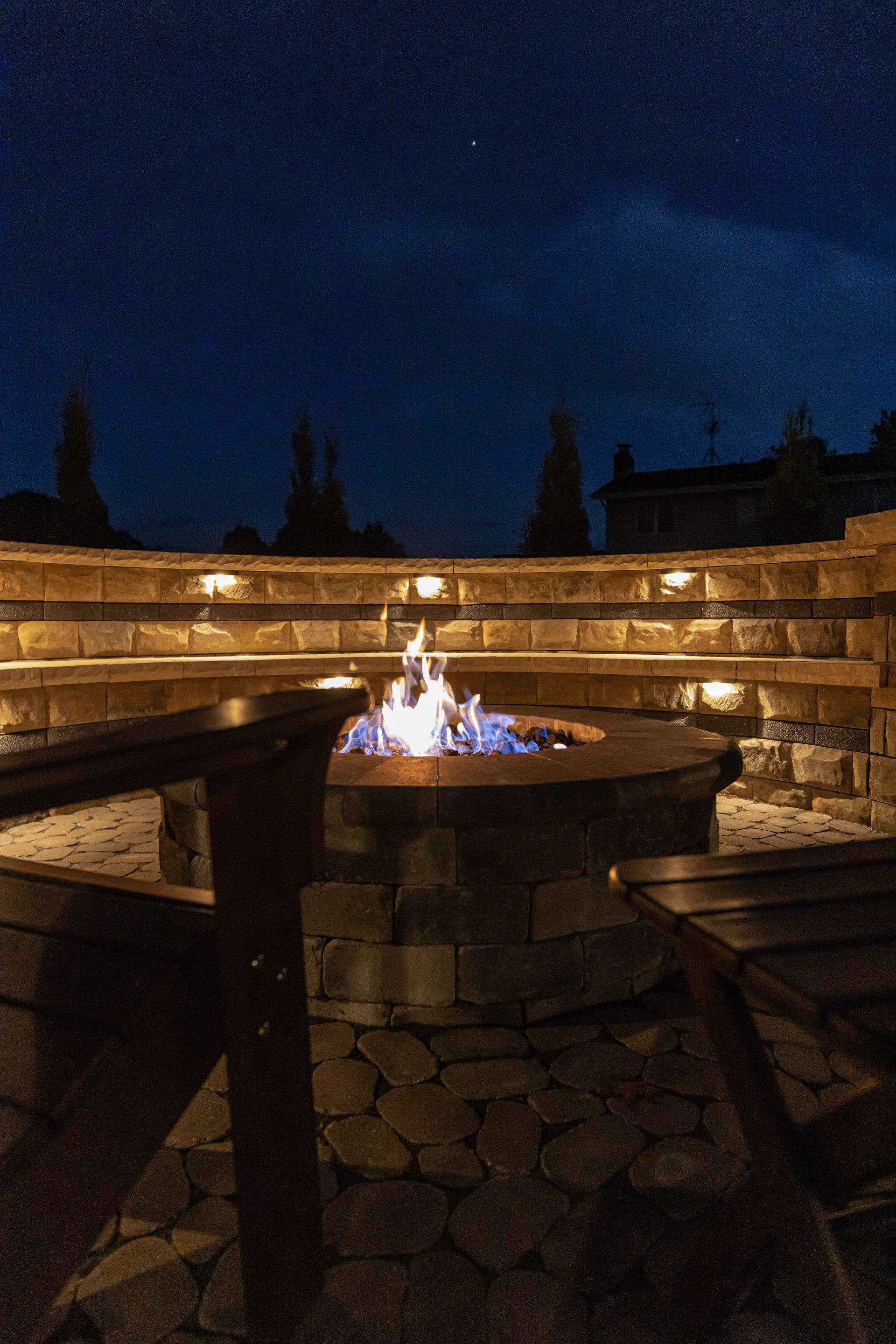 A nighttime shot of a stone fire pit burning surrounded by tiered seating, lit by overhead lights.