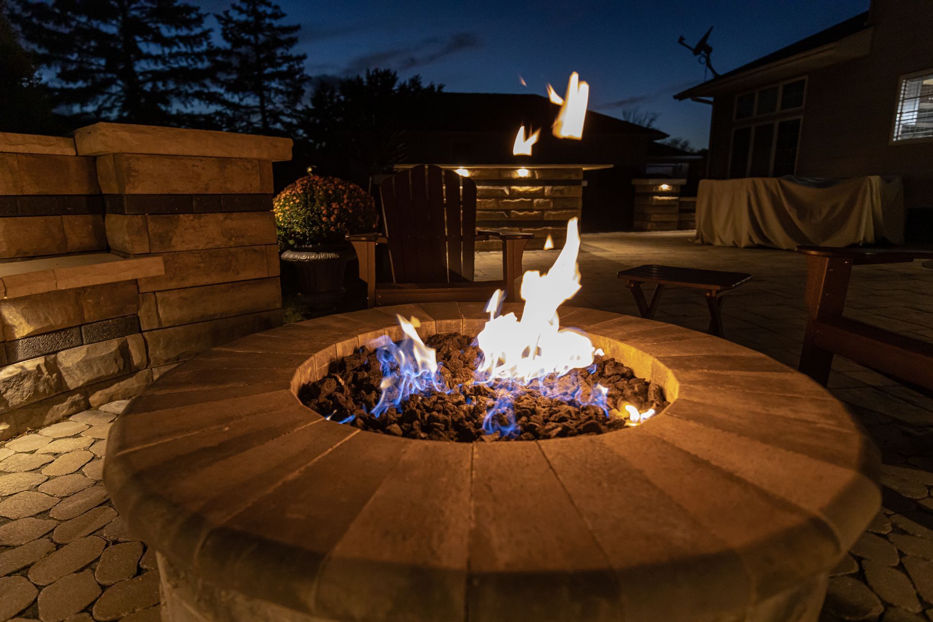 Fire pit ablaze in outdoor patio at dusk. Surrounded by stone, with seating nearby.