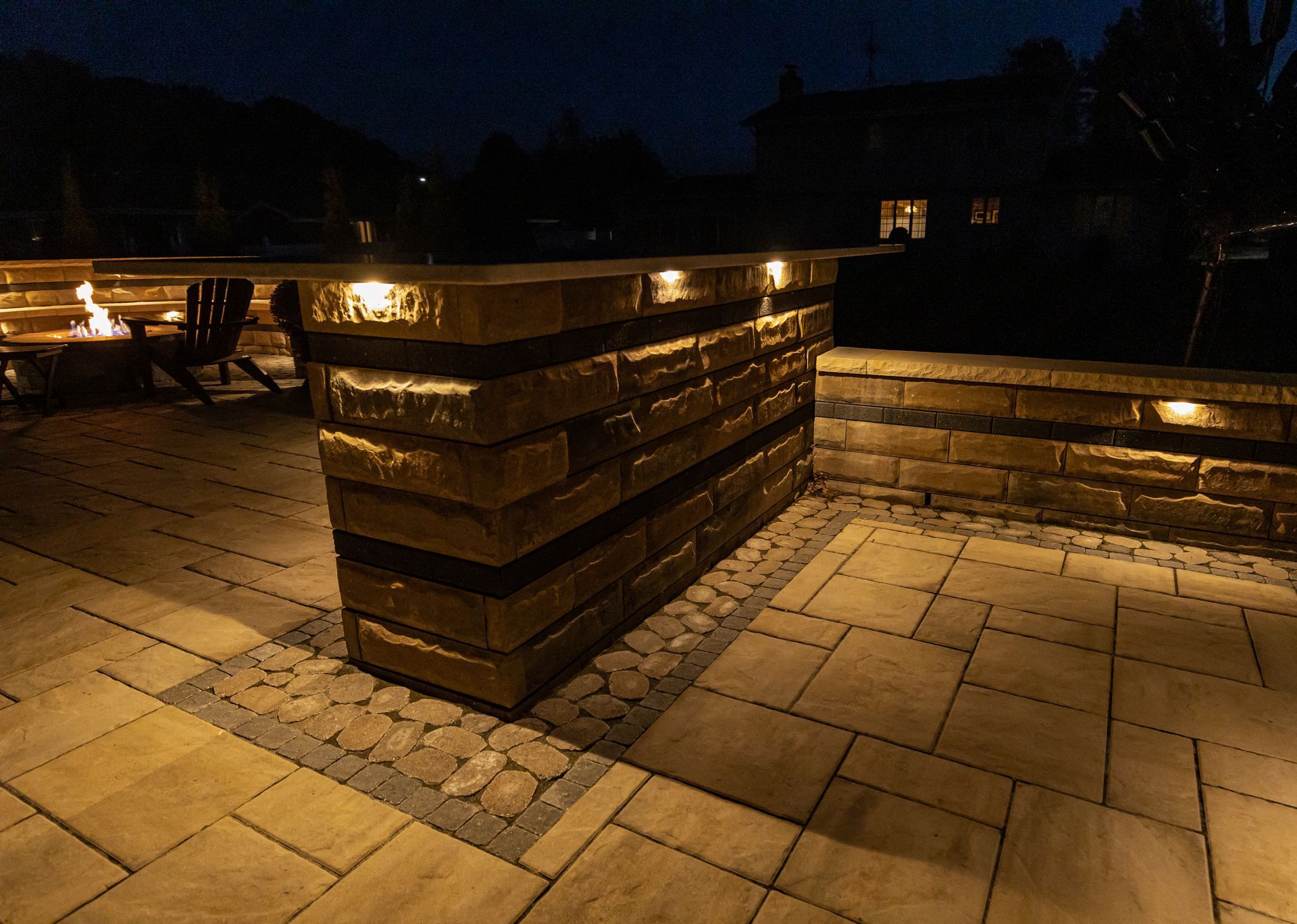 Outdoor stone bar with built-in lighting on a patio at night. Fire pit and house in the background.