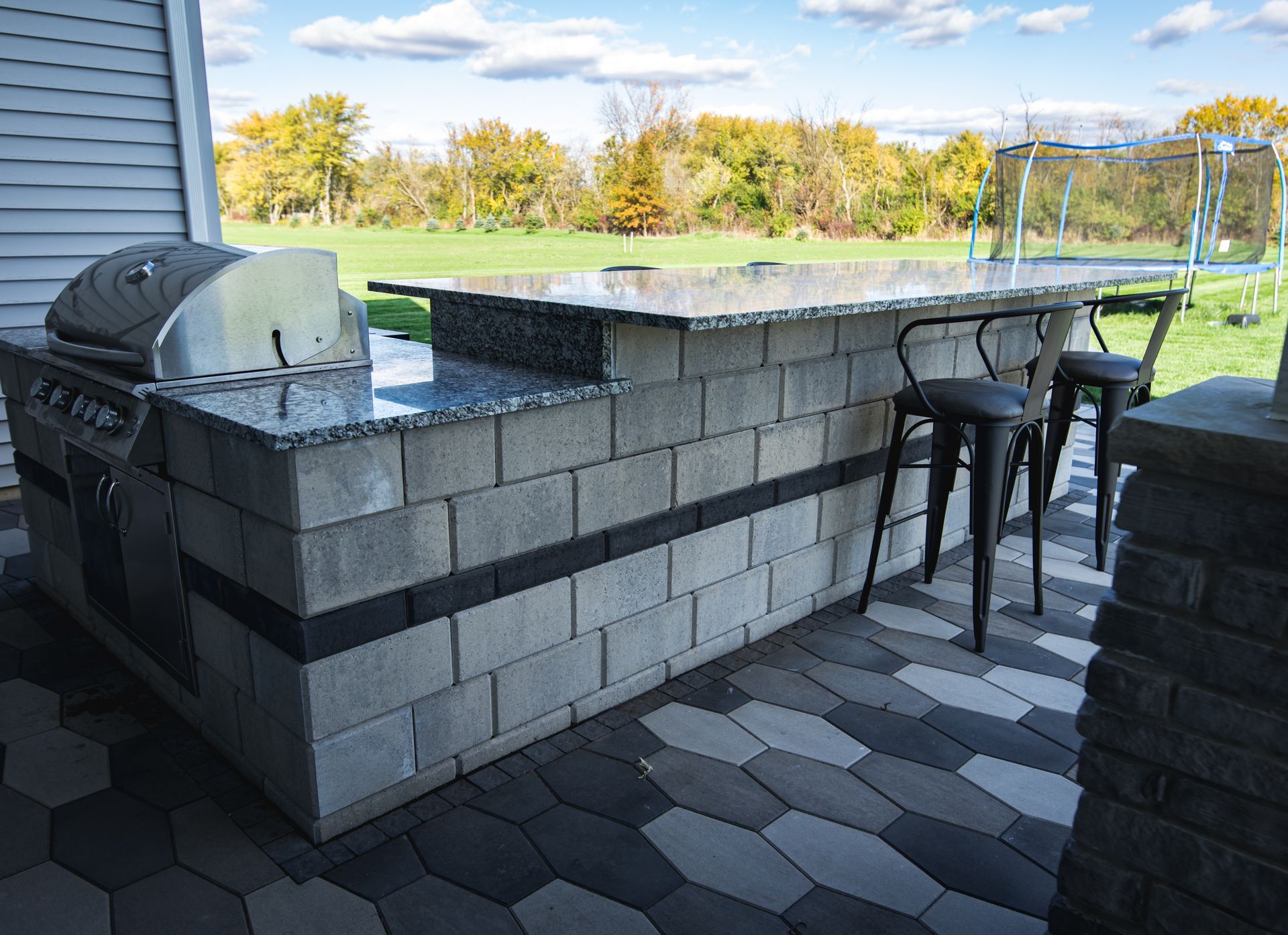 Outdoor kitchen with built-in grill and bar made of gray cinder blocks with black accents, topped with a granite countertop.