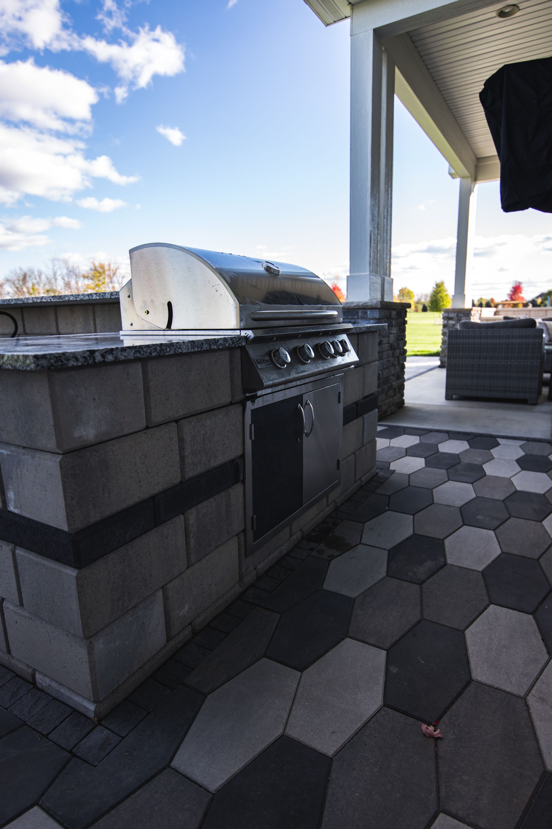Outdoor grilling station with stainless steel grill, on a stone block base, under a covered patio.