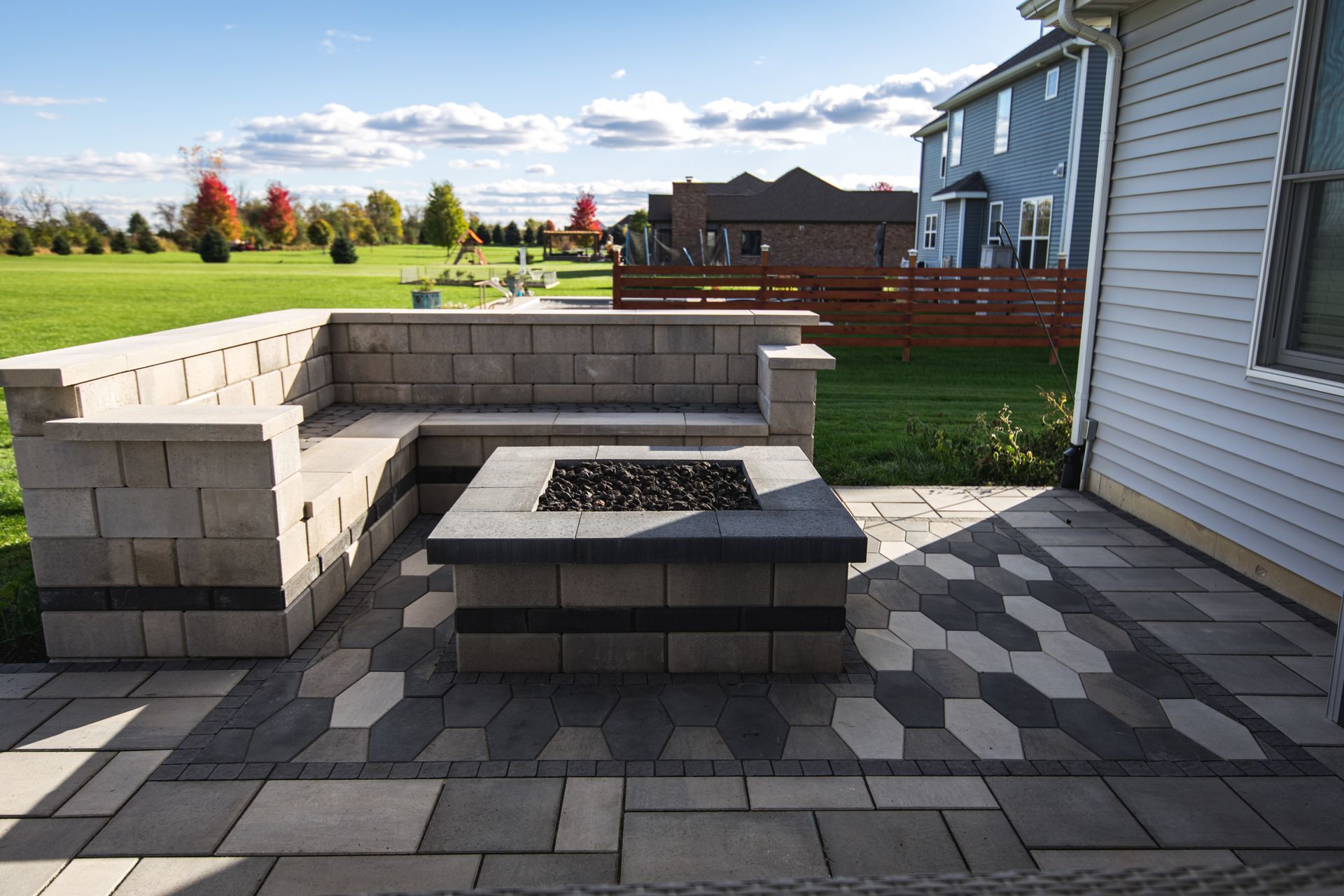 Backyard patio with built-in seating around a fire pit, surrounded by patterned paving stones and lawn.