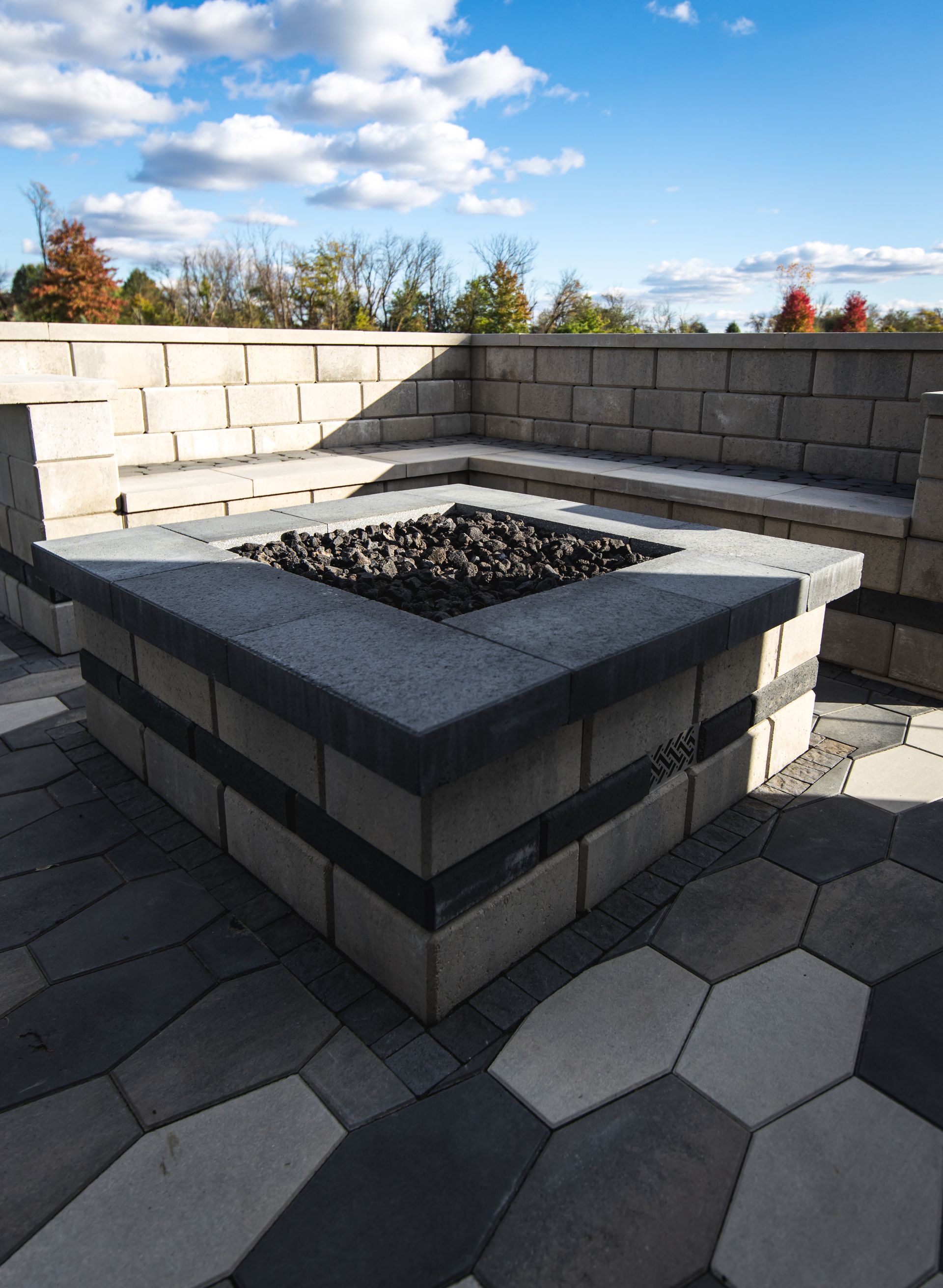 Fire pit and seating area made of stone blocks on a paved patio, with a blue sky background.