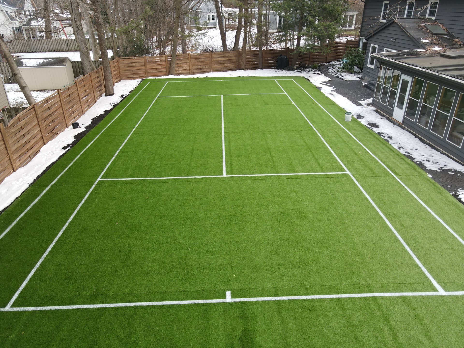 Artificial turf tennis court bordered by a wooden fence, with some snow.