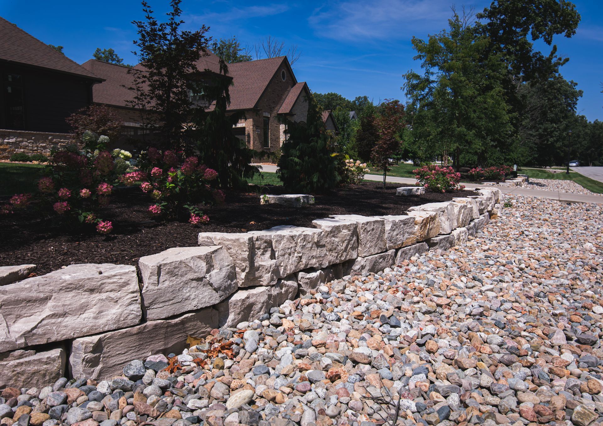 Low stone wall separating a gravel area from a landscaped flowerbed in front of houses under a blue sky.