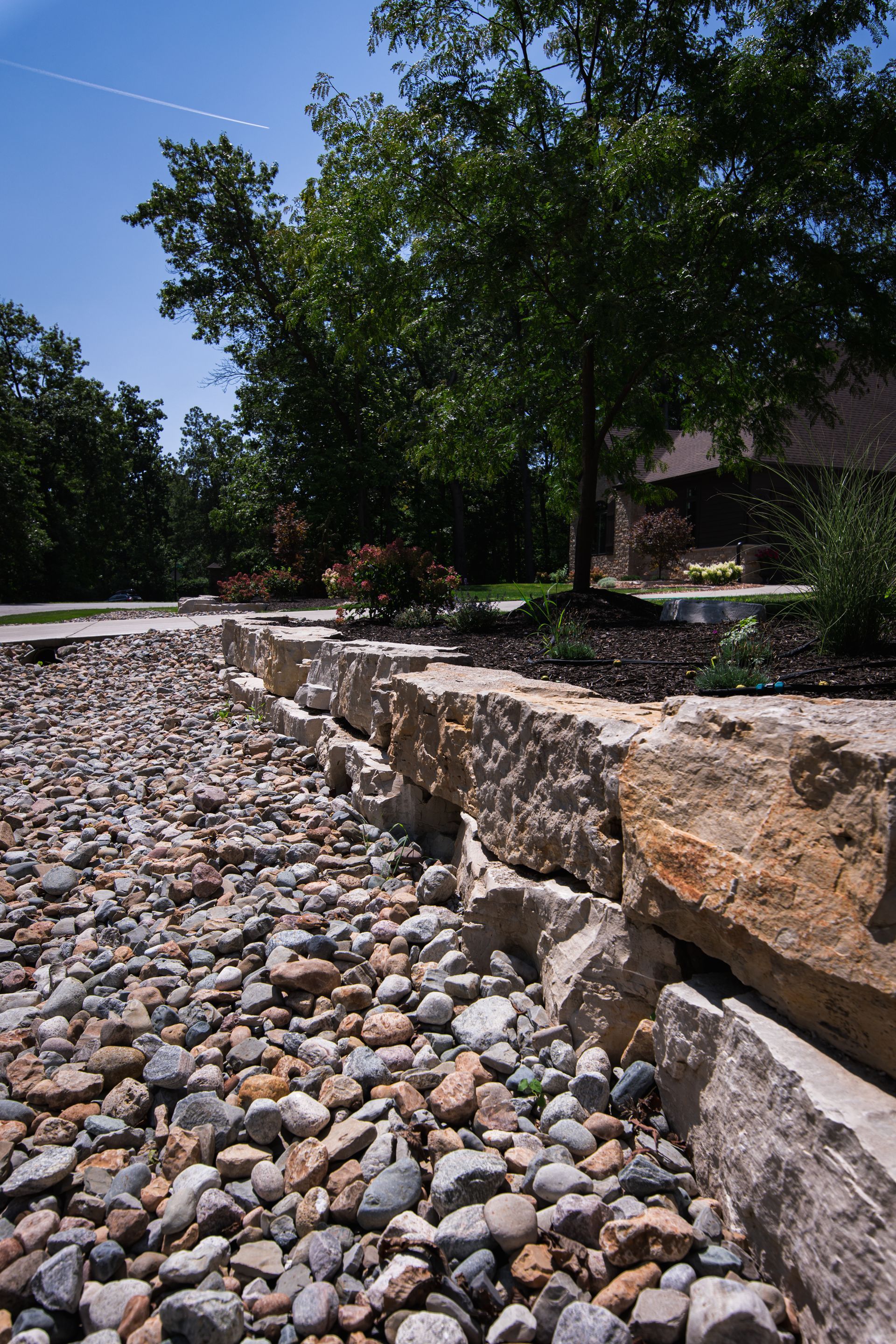 Stone retaining wall next to a bed of river rocks, with trees and a house in the background.