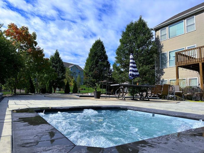Hot tub in a backyard with a house, trees, patio furniture, and blue sky.