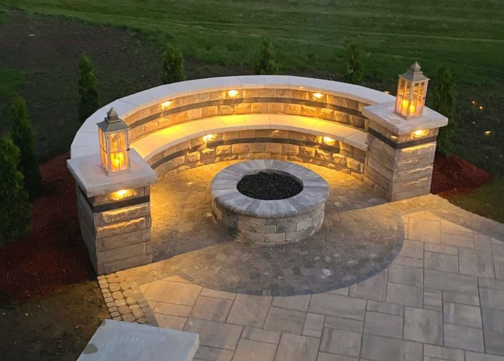 Fire pit ablaze in outdoor patio at dusk. Surrounded by stone, with seating nearby.
