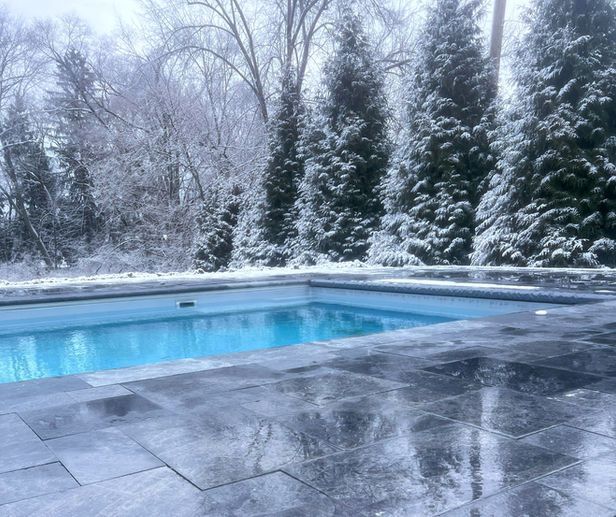 A pool with clear blue water surrounded by snow-covered patio and evergreens. Winter scene.