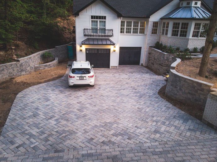 White SUV parked on a gray brick driveway leading to a two-story white house with black garage doors.