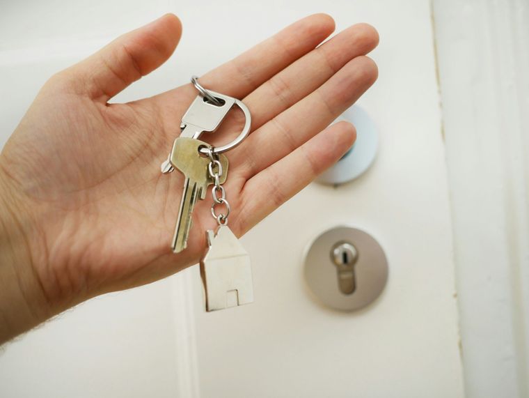 A hand holds a set of keys with a small house-shaped keychain in front of a white door.