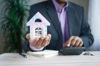 A person in a suit holds a small decorative house model while using a calculator at a desk with a notebook and pen.