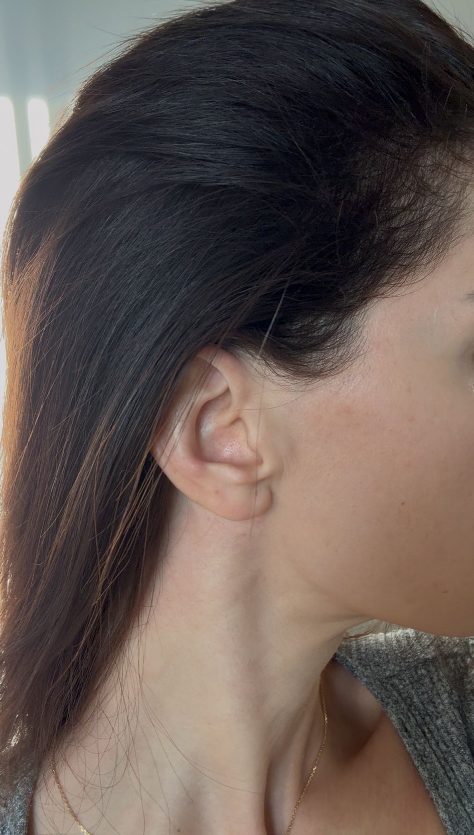 Profil einer Frau mit zurückgebundenem, dunklem Haar, Ohr und Hals sind sichtbar; sie trägt einen grauen Pullover.