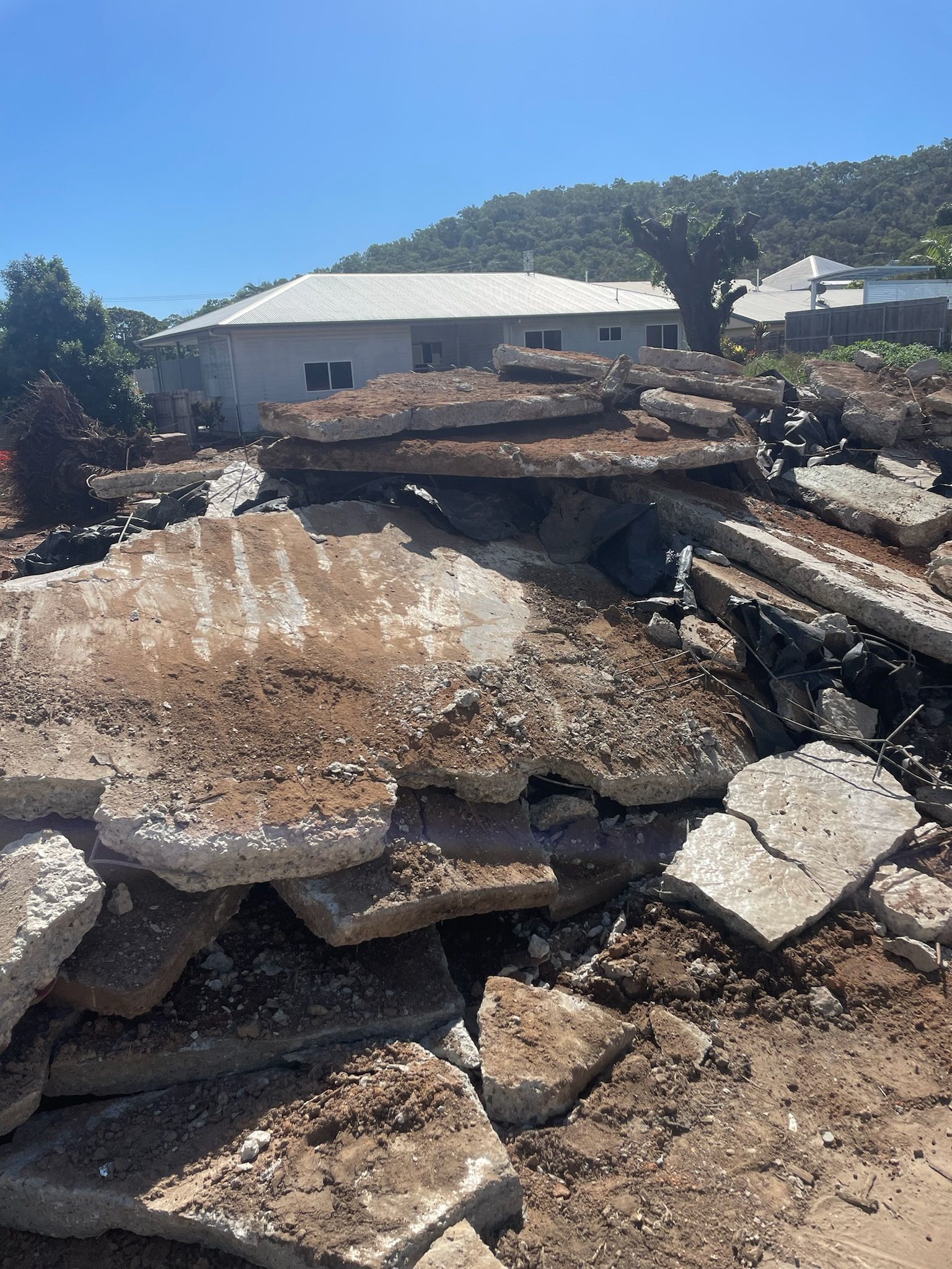 Pile of rubble in front of a house with a white roof on a sunny day.