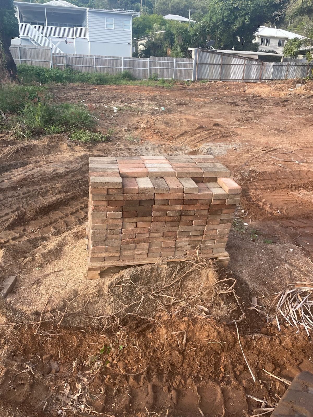 Stack of bricks on a wooden pallet, in a dirt lot. Houses and a fence are in the background.