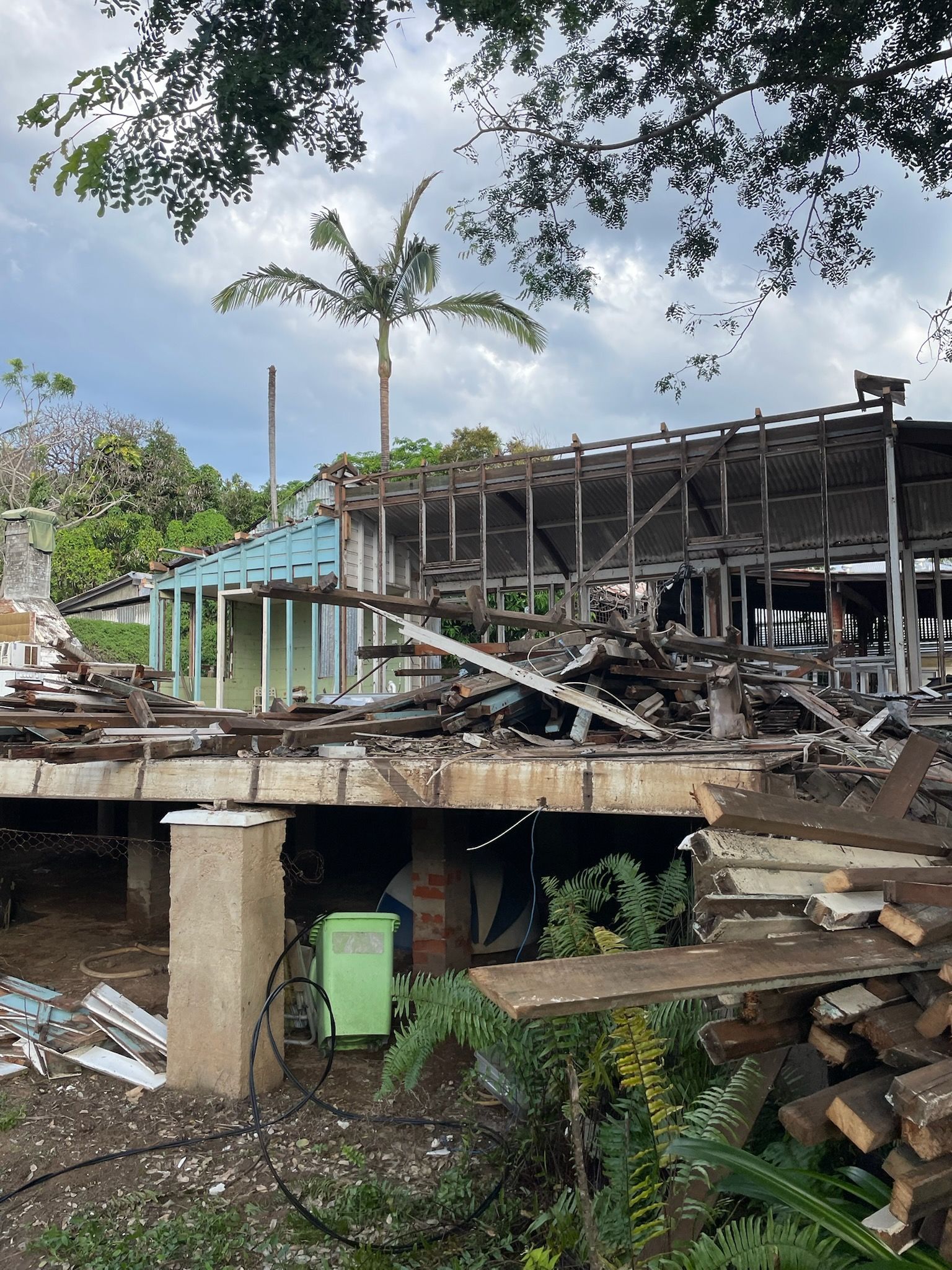 Building demolition site with exposed wood frame, debris, green bin, and palm tree. Cloudy sky.