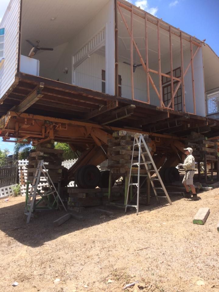 House raised on supports, construction in progress, man observing. White building, wood supports, blue sky.